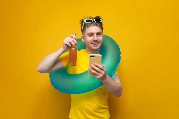 young guy on vacation in the summer drinks beer and uses a smartphone on a yellow background, a man with an inflatable swim ring looks at the phone and holds a bottle of drink