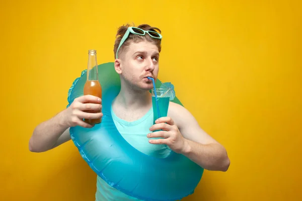 young guy on vacation in the summer holds a beer and a cocktail and dances on a yellow background, a man with an inflatable swim ring drinks refreshing drinks and relaxes