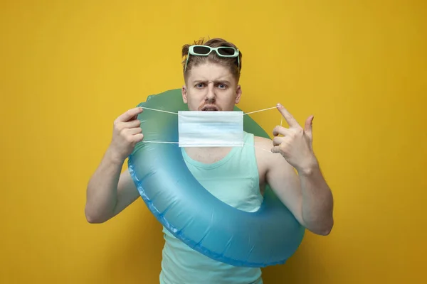young guy in the summer on vacation with an inflatable swim ring holds a medical mask and is surprised on a yellow background, a man is resting on the sea in a coronavirus pandemic