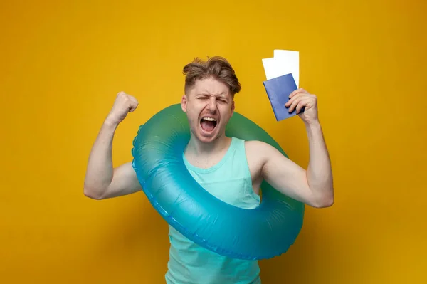 young guy tourist with an inflatable swim ring in the summer on vacation holds a passport with tickets and rejoice, a gesture of victory and success