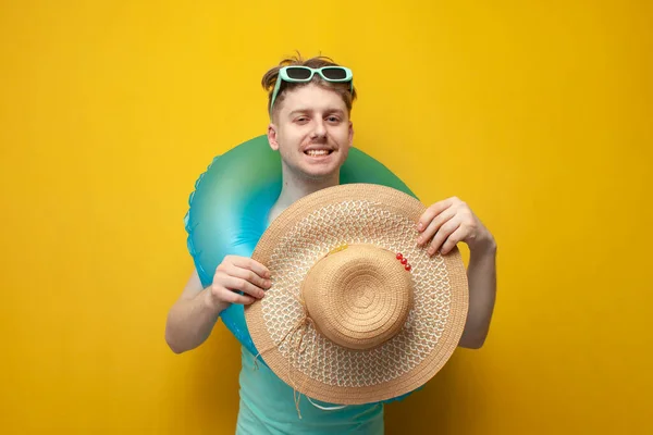 young guy in the summer on vacation with an inflatable swim ring holds a straw hat and smiles on a yellow background