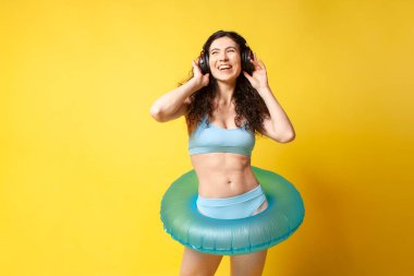 young curly slender brunette girl in a blue swimsuit in summer with an inflatable swim ring listens to music in headphones and dances, a woman on vacation on the beach enjoys the audio