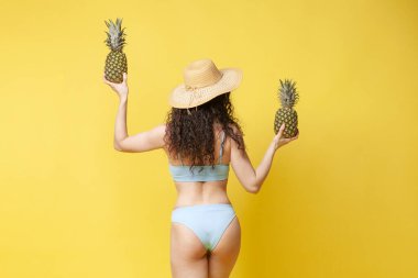 young curly brunette girl in a blue swimsuit dances with pineapples on a yellow background, a slender woman on the beach in summer with tropical fruits stands with her back