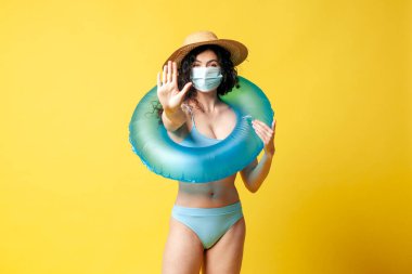 young brunette girl in a blue swimsuit with a swimming inflatable ring puts on a medical mask on a yellow background and shows a stop gesture