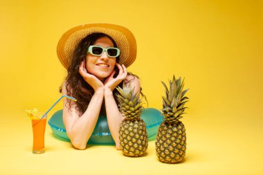 young girl in a blue sunglasses with an inflatable swimming ring sits on the beach and sunbathes in the summer, a woman with an orange cocktail on vacation at the sea on a yellow background