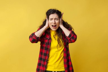 young curly girl covers her ears with her hands and screams on a yellow background, a student girl in stress is emotionally hysterical
