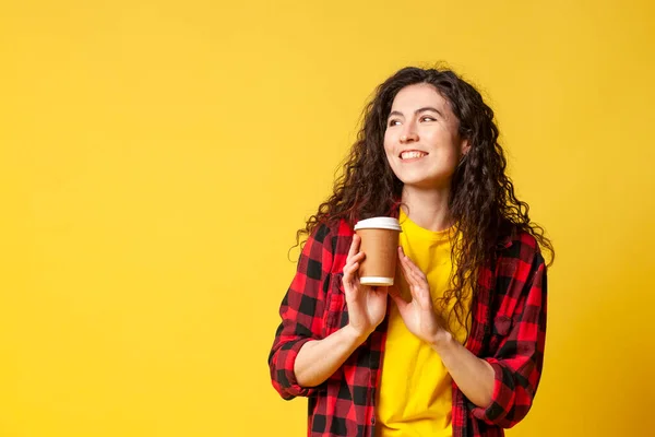 young curly brunette girl in a plaid shirt holds coffee on a yellow background and dreams, a student drinks coffee and looks at copy space