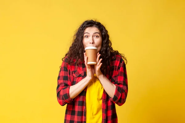 young shocked brunette girl in plaid shirt drinks coffee and shows surprise on yellow background, student shows charge of vivacity and energy from coffee