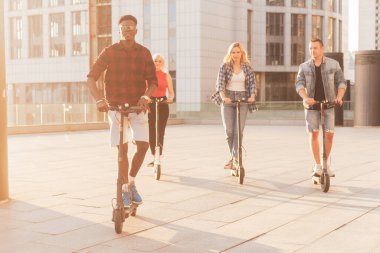 Irklar arası genç arkadaşlar şehirde elektrikli scooter kullanıyor, çok ırklı gençler elektrikli araç kullanıyor.