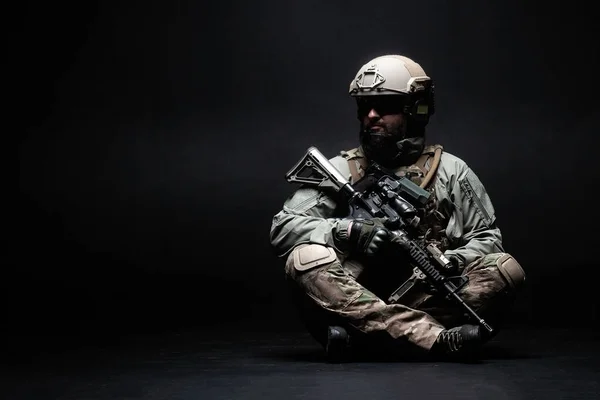 American soldier in a military uniform with a weapon sitting against a dark background and looking at the copy space, elite troops of America, American special forces