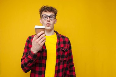 young curly guy in glasses holding coffee and wondering, student with a drink on a yellow background