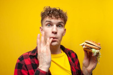 young surprised guy student eats a big tasty burger on a yellow background and licks the sauce on his finger, a man eats fast food and looks at the camera