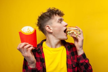 young curly guy holds french fries and bites a big tasty burger on a yellow background, student eats fast food