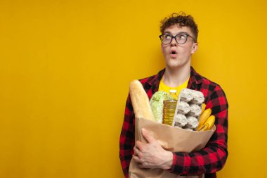 young shocked guy holds a package of groceries and is surprised on a yellow background, the buyer looks away at the copy space