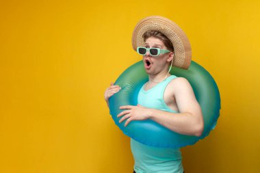 young guy in sunglasses and with inflatable swim ring in the summer on vacation shows surprise and looking on the copy space space on yellow background
