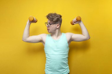 young non-athletic guy with glasses raises small dumbbells on a yellow background and shows surprise, a nerd is training and doing sports