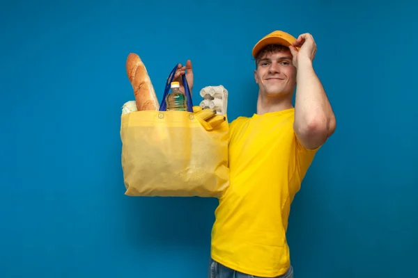 young food delivery man holds a yellow eco bag on a blue background, a courier in a yellow uniform delivers a cloth non-plastic bag full of groceries