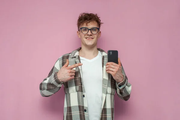 young curly guy in glasses holds a smartphone and points a finger at the phone on a pink background, a man with a mobile phone smiles