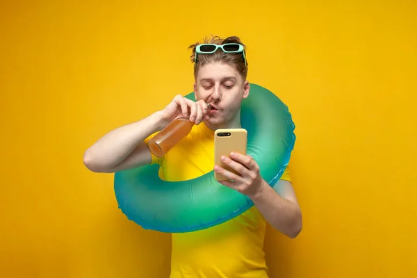 young guy on vacation in the summer drinks beer and uses a smartphone on a yellow background, a man with an inflatable swim ring looks at the phone and holds a bottle of drink