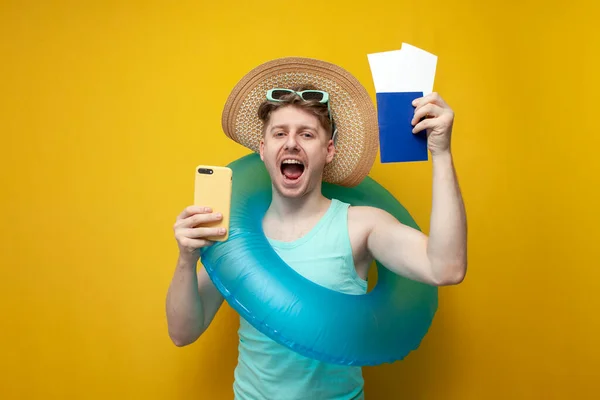 young guy tourist with an inflatable swim ring in the summer on vacation holds a passport with tickets and a smartphone and rejoices on a yellow background, the concept of travel and buying tickets