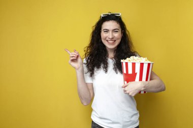 young brunette girl viewer in 3d glasses and white t-shirt holds large bucket of popcorn and shows her hand aside on copy space