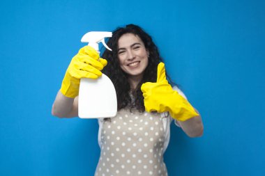 young girl cleaner in uniform holds a spray and shows a like on a blue background, a woman housekeeper in an apron and gloves holds cleaning products