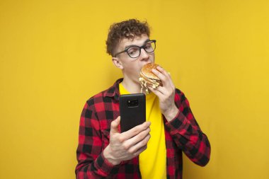 young curly guy in glasses holds a big burger and uses a smartphone, a student eats fast food and looks into the phone on a yellow background