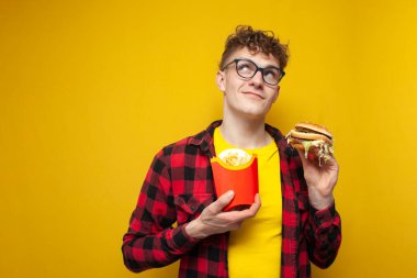 young curly guy with glasses holding a burger and fries thinks and looks up, a student with fast food dreams on a yellow background