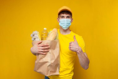 Sarı üniformalı ve tıbbi maskeli genç kurye sarı arka planda bir paket erzak tutuyor. Coronavirus salgınında yemek dağıtan adam.