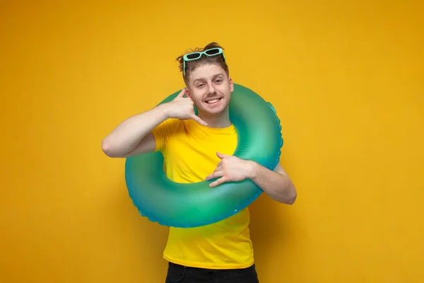 call me. young guy tourist with an inflatable swim ring shows a call gesture on a yellow background, the concept of a summer vacation at sea