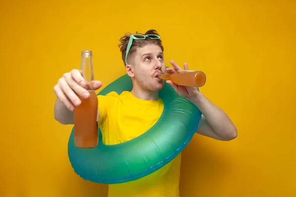 young happy guy in the summer on vacation drinks beer and offers a drink on a yellow background, a man with an inflatable swim ring is resting on the sea with alcoholic drinks