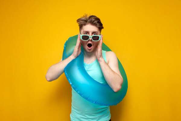 молодой шокированный парень в солнечных очках и с надувным плавательным кольцом летом на отдыхе показывает сюрприз на желтом фоне