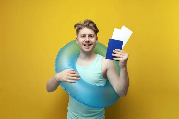 young guy tourist with an inflatable swim ring in the summer on vacation holds a passport with tickets and smiles on a yellow background, the concept of traveling to the sea