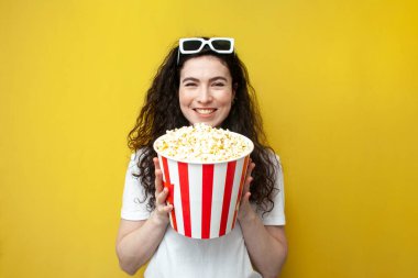 young curly cheerful woman in glasses holds popcorn and smiles on yellow background, the concept of watching movie, girl cinema viewer