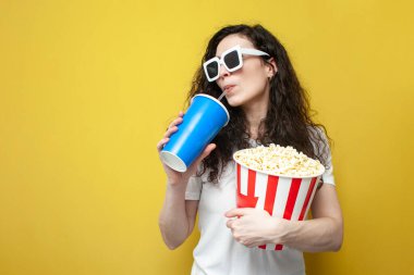 young brunette girl viewer in 3d glasses and a white t-shirt holds a large bucket of popcorn and drinks soda on yellow isolated background