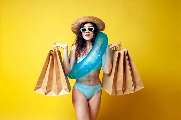 young attractive brunette girl in a blue swimsuit with a swimming inflatable ring holds packages with purchases and goes shopping on a yellow background, a woman shopper in the summer on vacation
