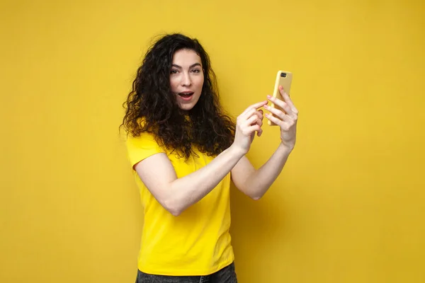 young curly woman in yellow t-shirt holds smartphone and presses her finger on the screen, concept of online shopping, the girl uses the phone on yellow background