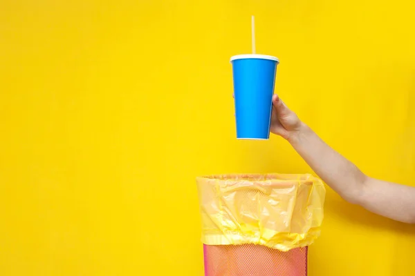hand throws plastic cup into trash can on yellow background, ecology concept, garbage falls into trash can