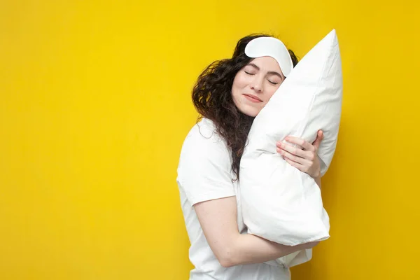 young woman in sleep mask hugs white soft pillow on yellow background and sleeps, the concept of healthy sleep and dreams
