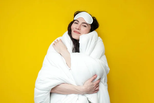 young woman in sleep mask covers herself with white fluffy soft blanket and smiles on yellow background, the concept of healthy sleep and bed linen, girl hugs rug and wants to sleep