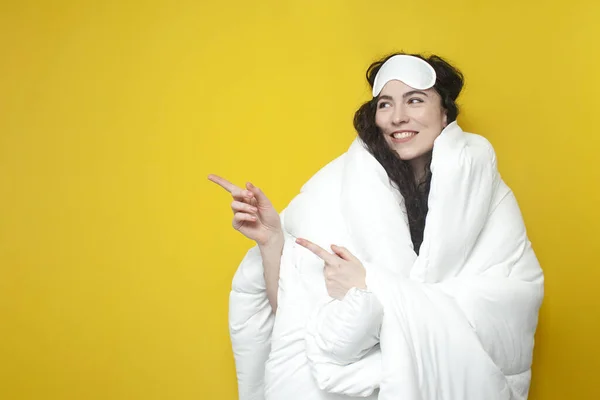 young woman in sleep mask covers herself with white fluffy soft blanket and shows on the copy space, concept of healthy sleep, girl hugs the carpet and advertises place for text