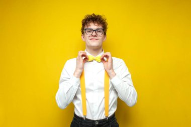 young curly guy in shirt and suspenders straightens his bow tie on yellow background, smart nerd in glasses on yellow isolated background