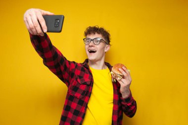young curly guy holds a big burger and takes a selfie on a smartphone, a student photographs himself with fast food on a yellow background