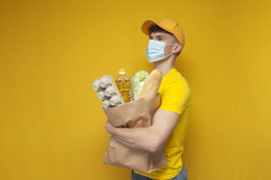 Sarı üniformalı ve tıbbi maskeli genç kurye sarı arka planda bir paket erzak tutuyor. Coronavirus salgınında yemek dağıtan adam.