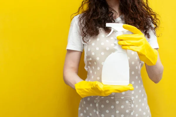 young girl cleaner in apron and gloves holds cleaning agent on yellow background, empty cleaning spray in the hands of housewife, mock-up