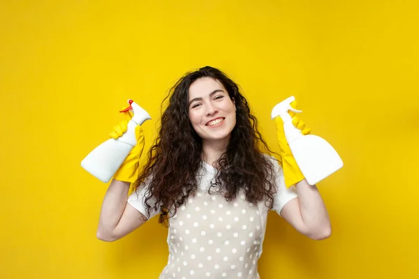 young girl cleaner in apron and gloves holds cleaning agent on yellow background, girl housewife with chemistry for cleaning