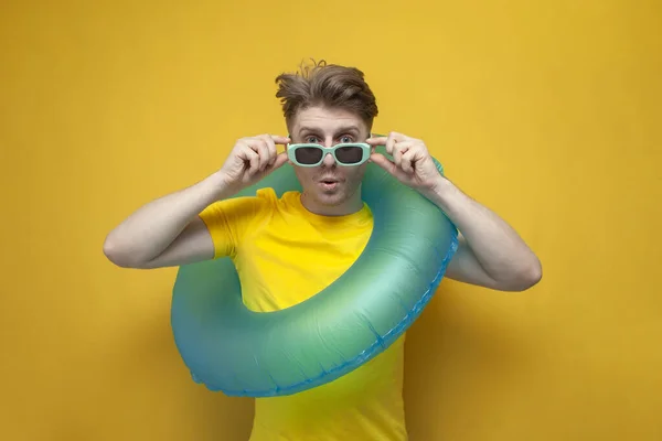 surprised young guy on vacation with swim ring holding blue goggles on yellow background, sea travel concept