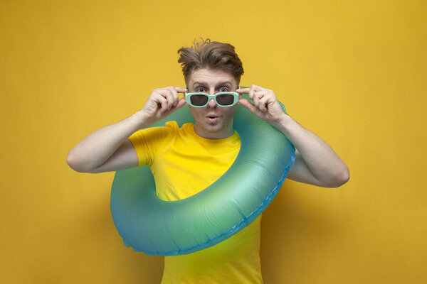 surprised young guy on vacation with swimming ring holding blue goggles on yellow background, sea travel concept