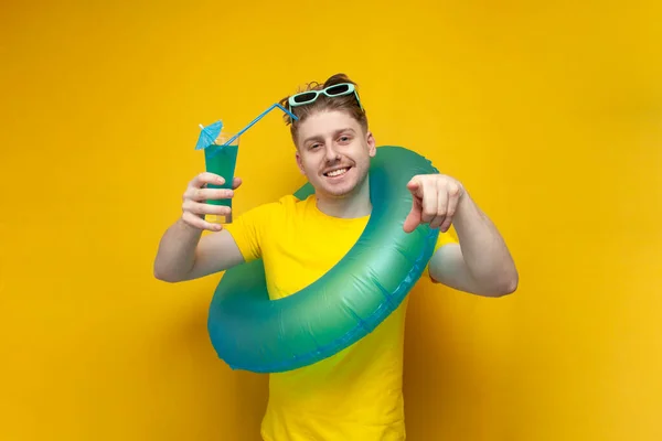 I choose you. a young guy in the summer on vacation holds a swimming inflatable ring and drinks a refreshing blue cocktail on a yellow background, man with summer drink points his finger at the camera