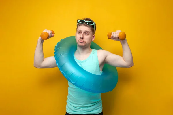 young guy with an inflatable swim ring in the summer on vacation goes in for sports and raises small dumbbells on a yellow background